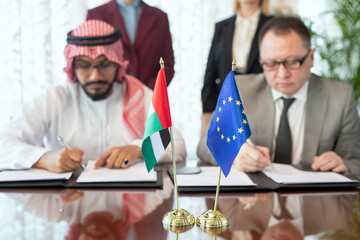 A person in a suit and tie and Arabian businessman signing documents by table