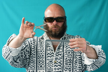 A guy with a bald head and a lush beard holds scissors and a comb in his hands. Barber hairdresser. A man with glasses and a shirt.  Stylish bearded man. Portrait                            