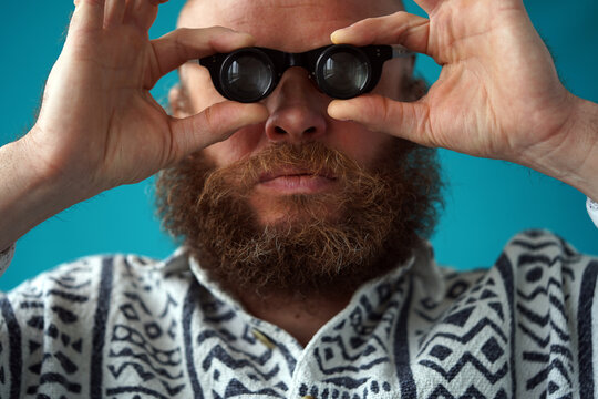 A man in glasses with a mustache and a beard holds binoculars in his hands. Discovery idea. Portrait. A bald-headed guy in a colorful shirt. Bearded