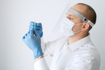 Naklejka premium Vaccination and immunization. A man wearing gloves, a mask and a protective shield holds an injection syringe, shaking it from air bubbles. Flu vaccine, coronavirus vaccine. A doctor in a white coat