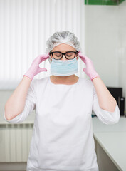 tired female doctor wearing glasses and a white uniform puts on a disposable protective mask in the clinic office