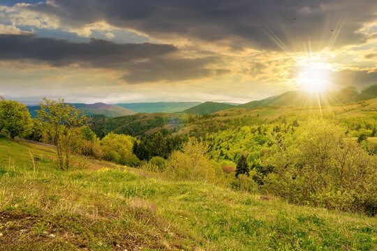 rural mountain landscape in spring at sunset. grass and trees on hills rolling through the green valley in to the distant ridge beneath a cloudy sky in evening light
