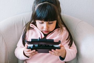 Little black haired girl wearing a pink sweater playing a video game with a portable video console on a sofa