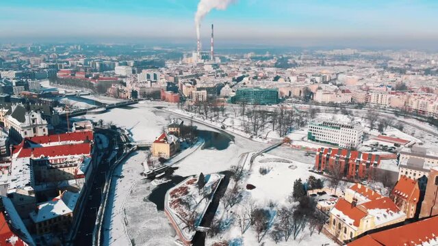 Drone footage of the industrial area of the city of Wroclaw, Poland. Harmful smoke coming out of the chimneys of the power plant polluting the blue sky. Winter season. The city covered with snow.