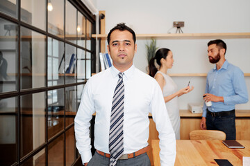 Portrait of serious Indian entrepreneur in white shirt looking at camera, his coworker talking in background