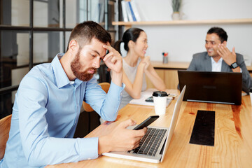 Bored young businessman sitting at table in meeting room and checking social media on smartphone