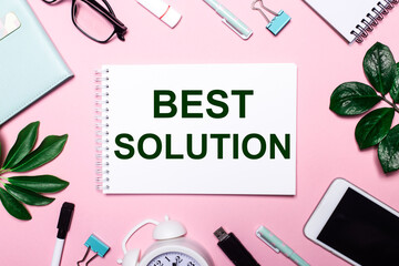 BEST SOLUTION is written in a white notebook on a pink background surrounded by business accessories and green leaves.