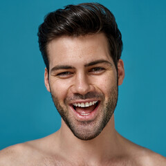 Beauty portrait of joyful naked brunette man with fresh smooth skin and perfect smile looking at camera, posing isolated over blue background