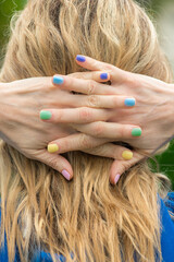 Woman head from behind, blond hair, rainbow nail polish manicure, all fingers crossed.