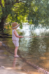 Photo of little blond caucasian barefoot boy in straw hat fishing on the river holding handmade wooden fishing rod. Hobbies and leisure activity theme.