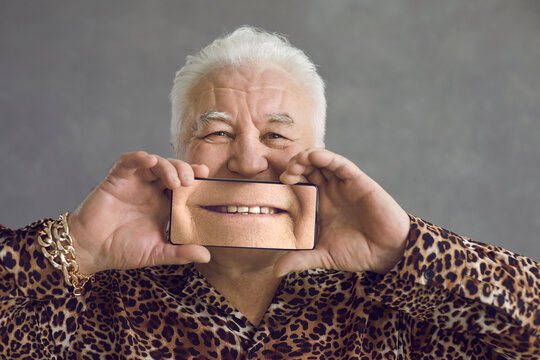 Portrait of funny positive fat white haired senior pensioner in leopard shirt and bling gold chain bracelet covering face with modern cell phone with magnified perfect smile image. Dental care concept