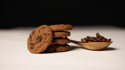 A stack of chocolate cookies beside a spoon of brown coffee beans in dark background