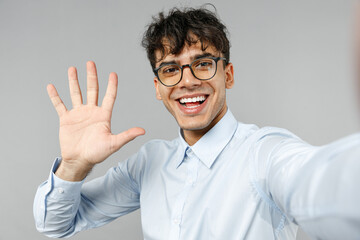 Close up young smiling happy employee business latin man 20s corporate lawyer in classic white shirt glasses doing selfie shot on mobile phone waving hand greeting isolated on grey background studio