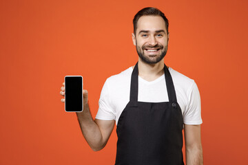 Young man barista bartender barman employee in black apron white t-shirt work in coffee shop show hold mobile phone blank screen workspace area isolated on orange background. Small business startup.