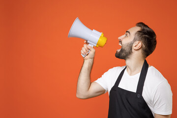 Side view young man barista bartender barman employee wearing black apron white t-shirt work in coffee shop scream in megaphone shout spread hands isolated on orange background Small business startup