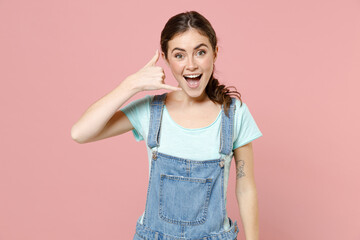 Young excited cheerful caucasian woman in trendy denim clothes blue t-shirt doing phone gesture like says call me back isolated on pastel pink background studio portrait. People lifestyle concept.