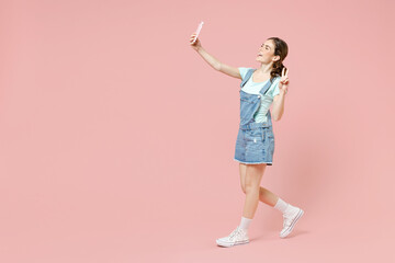 Fototapeta na wymiar Young smiling happy caucasian woman 20s wearing trendy denim clothes blue t-shirt doing selfie shot on mobile phone showing victory v-sign gesture isolated on pastel pink background studio portrait