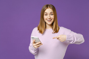 Naklejka premium Young surprised happy student caucasian woman 20s in purple knitted sweater point index finger on mobile cell phone chat online isolated on violet background studio portrait. People lifestyle concept.