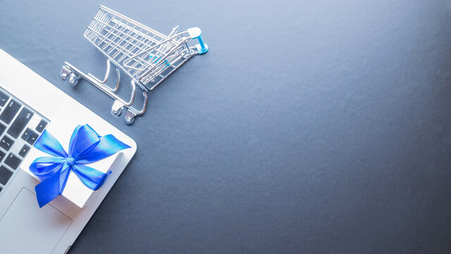 Online shopping flat. Laptop computer, shopping trolley and white gift with blue ribbon on dark background. Internet purchase, online shop concept. Website retail business.