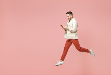 Fototapeta na wymiar Full length young fun trendy fashionable caucasian man 20s in jacket white t-shirt jump high using mobile cell phone chatting online in social network isolated on pastel pink color background studio.