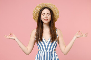 Young woman in summer clothes striped dress straw hat hold spread hands in yoga om gesture relax meditate try to calm down isolated on pastel pink background studio portrait. People lifestyle concept.