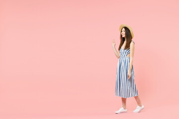 Full length young fun smiling happy caucasian woman in summer clothes striped dress straw hat walk going waving hand greet isolated on pastel pink background studio portrait. People lifestyle concept.