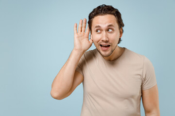 Fototapeta premium Young curious nosy excited expressive man 20s wear casual basic beige t-shirt try to hear you overhear listening intently isolated on pastel blue background studio portrait. People lifestyle concept.