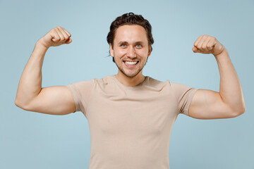 Young strong sporty fitness man in casual basic beige t-shirt showing biceps muscles on hand demonstrating strength power isolated on pastel blue background studio portrait. People lifestyle concept.
