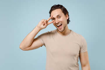 Fototapeta premium Young fun smiling happy friendly caucasian man wearing casual basic beige t-shirt cover eye with victory v-sign gesture isolated on pastel blue background studio portrait. People lifestyle concept.