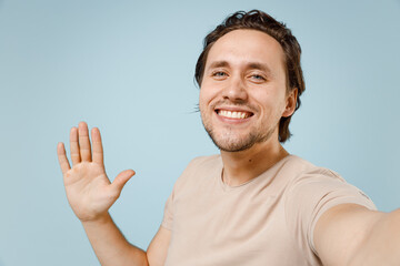 Close up young smiling man 20s wearing casual basic beige t-shirt doing selfie shot on mobile phone waving hand greeting isolated on pastel blue background studio portrait. People lifestyle concept.