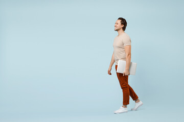 Fototapeta na wymiar Full length young smiling happy successful caucasian man in casual basic beige t-shirt hold closed laptop pc computer walk isolated on pastel blue background studio portrait. People lifestyle concept.