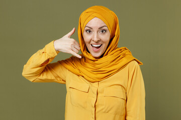 Friendly young arabian asian muslim woman in abaya hijab yellow clothes do phone gesture like says call me back isolated on olive green background. People uae middle eastern islam religious concept