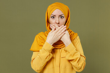 Shocked scared astonished young arabian asian muslim woman in abaya hijab yellow clothes cover mouth with hands isolated on olive green background People uae middle eastern islam religious concept