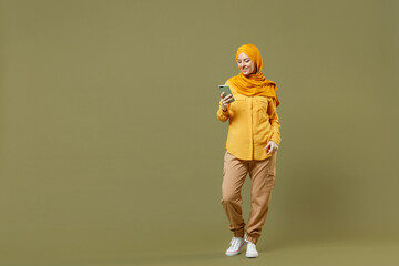 Full length young arabian asian muslim woman in abaya hijab yellow clothes hold in hand using mobile cell phone isolated on olive green background studio portrait. People uae islam religious concept.