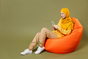 Full length young arabian asian muslim woman in abaya hijab yellow clothes hold mobile cell phone sit in bean bag chair isolated on olive green background studio. People uae islam religious concept.