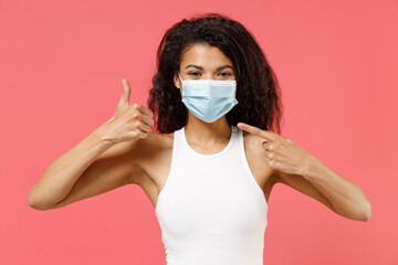 Young african american woman in white tank shirt point index finger on sterile face mask ppe coronavirus virus covid-19 flu on lockdown quarantine show thumb up gesture isolated on pink background