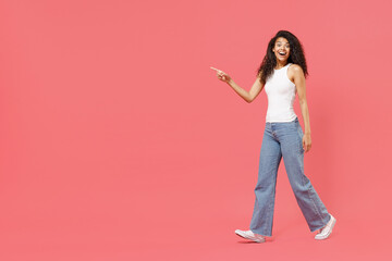 Fototapeta na wymiar Full length young fun smiling happy friendly positive african american woman 20s wear casual white tank shirt walk go point index finger aside on copy space area mock up isolated on pink background