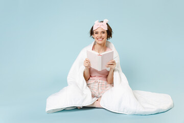 Young fun smiling woman 20s in pajamas jam sleep eye mask rest at home sit wrap cover under blanket duvet read hold book novel diary isolated on pastel blue background Good mood night bedtime concept
