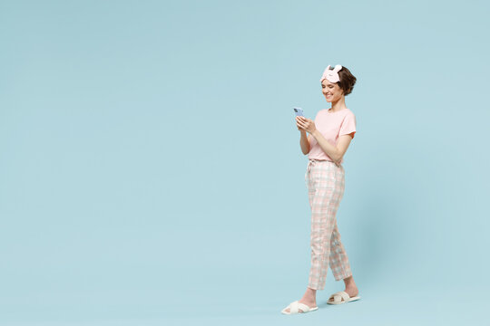 Full length side view happy young woman in pajamas jam sleep eye mask rest relaxing at home hold mobile cell phone walking going isolated on pastel blue background. Good mood night bedtime concept