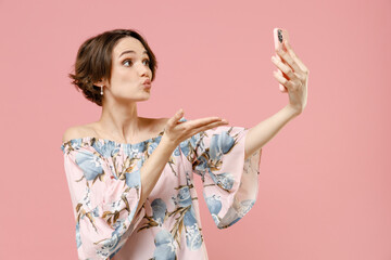 Young friendly happy woman 20s with short hairdo wear stylish blouse doing selfie shot on mobile phone post photo on social network blowing air kiss isolated on pastel pink color background studio