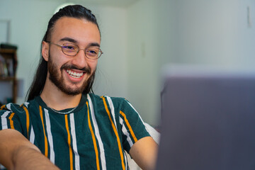 young latin long haired man laughing happily, while using the computer at home to study, or work freelance