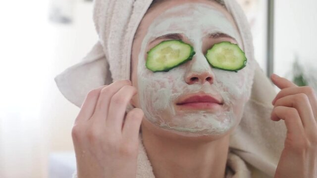 Young pretty woman with cosmetic carbo detox clay light green mask put slices of cucumber, covering eyes and relaxing. Girl takes care of oily skin complexion. Beauty treatment. Skincare