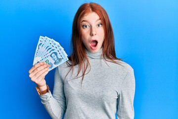 Young red head girl holding 10 yuan chinese banknotes scared and amazed with open mouth for...