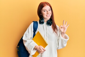 Young red head girl wearing student backpack and holding book doing ok sign with fingers, smiling friendly gesturing excellent symbol