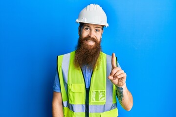 Redhead man with long beard wearing safety helmet and reflective jacket showing and pointing up with finger number one while smiling confident and happy.