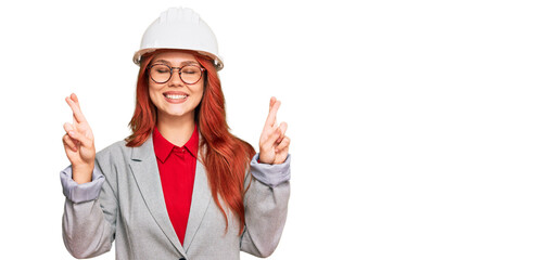 Young redhead woman wearing architect hardhat gesturing finger crossed smiling with hope and eyes closed. luck and superstitious concept.
