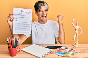 Young hispanic man artist holding passed exam sitting on the table screaming proud, celebrating victory and success very excited with raised arm