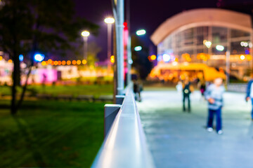 Buildings near the road. People go on the road. Night in the city. Trees on the lawn. Close up view from the handrail on the sidewalk level.