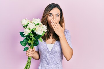 Young hispanic girl holding flowers covering mouth with hand, shocked and afraid for mistake....