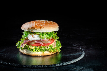 Big tasty burger with grilled meat, cheese, tomato, salad, onion rings and fried eggs on a black stone background